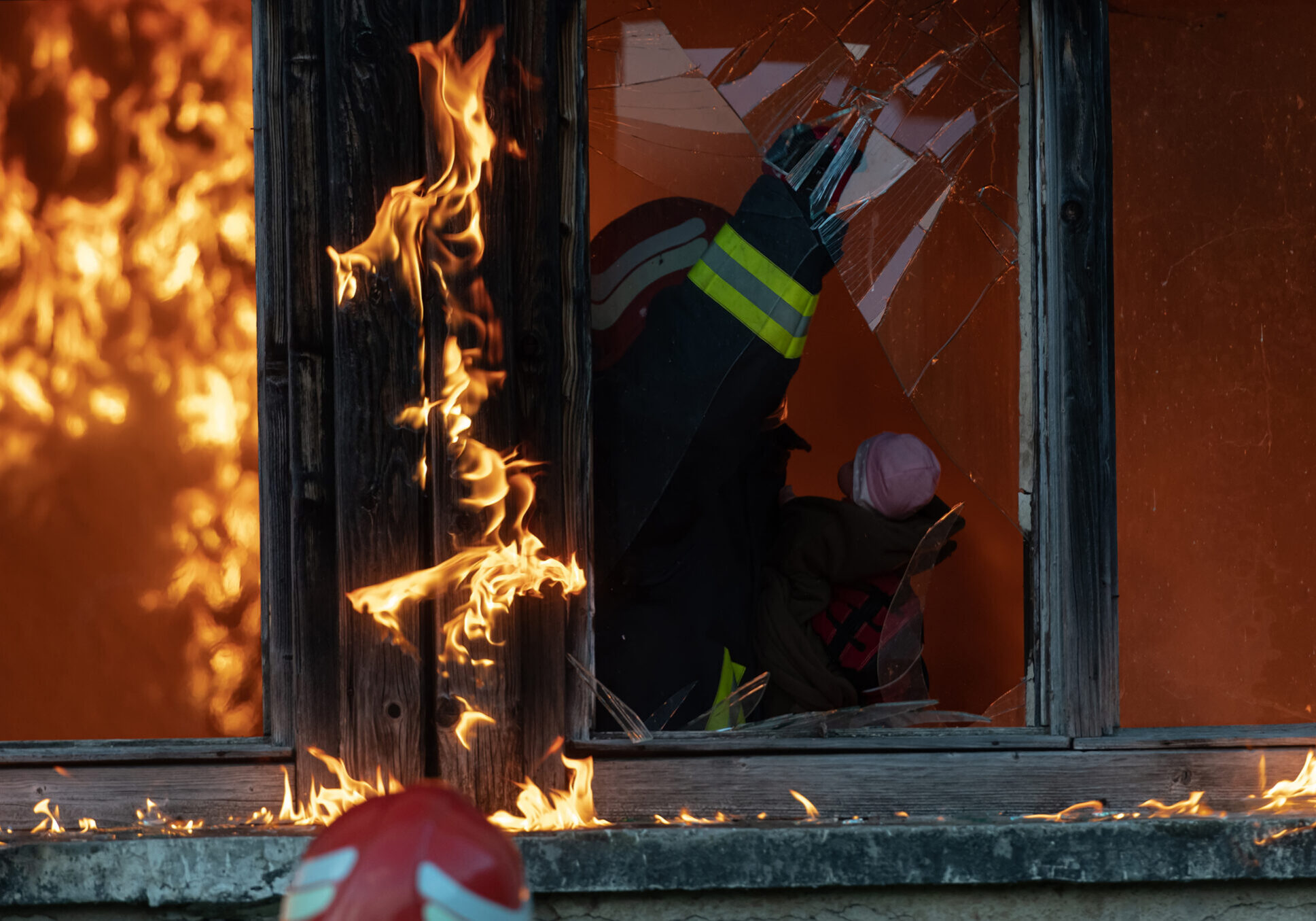 Firefighter battling flames and smoke damage in a window during a fire.